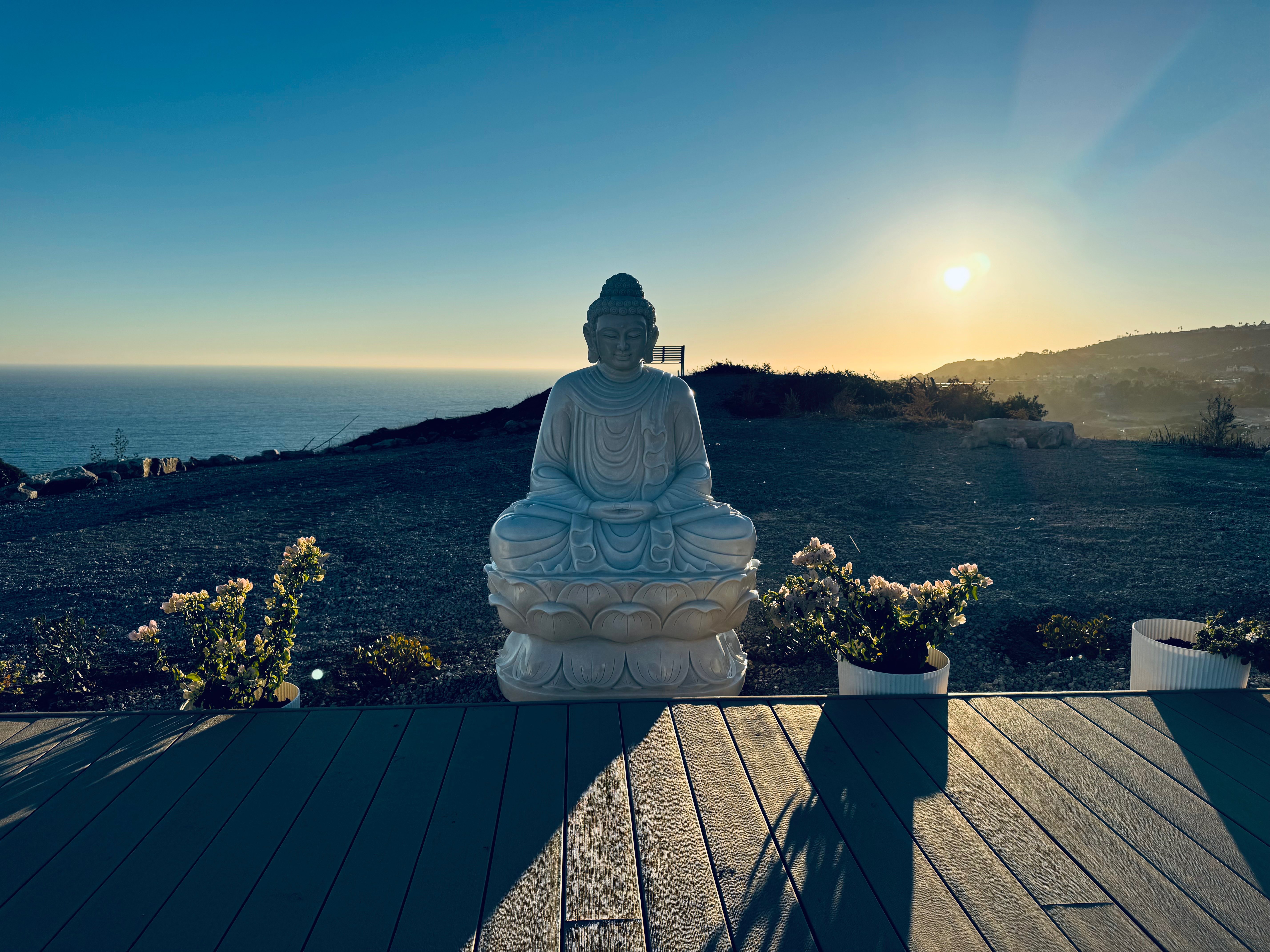 Golden hour meditation scene with Buddha statue on wooden deck overlooking Pacific Ocean