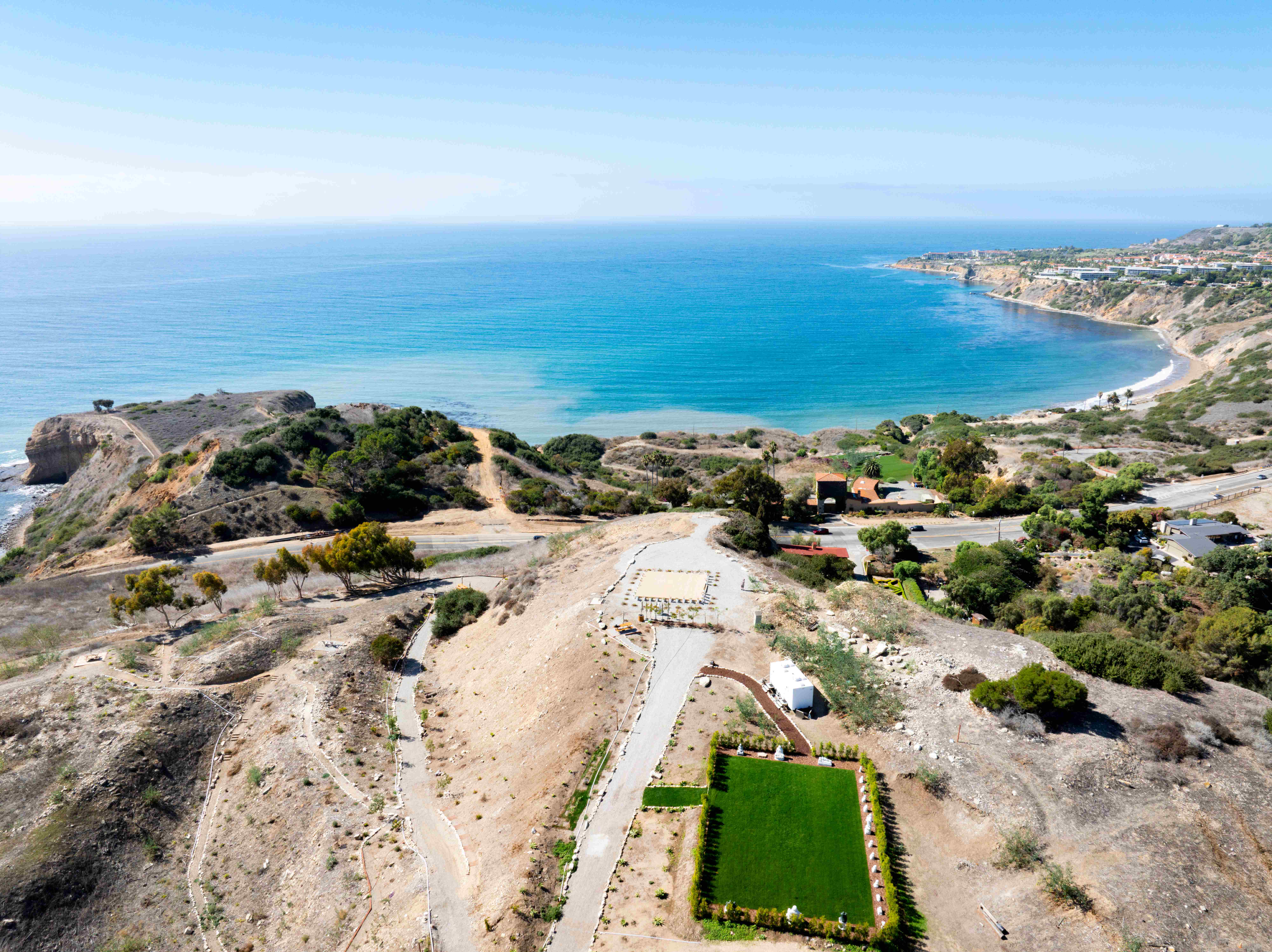 Stunning aerial view of Palos Verdes coastline with turquoise ocean, rocky promontory, and Infinite Hill property with Plaza Sunshine in foreground