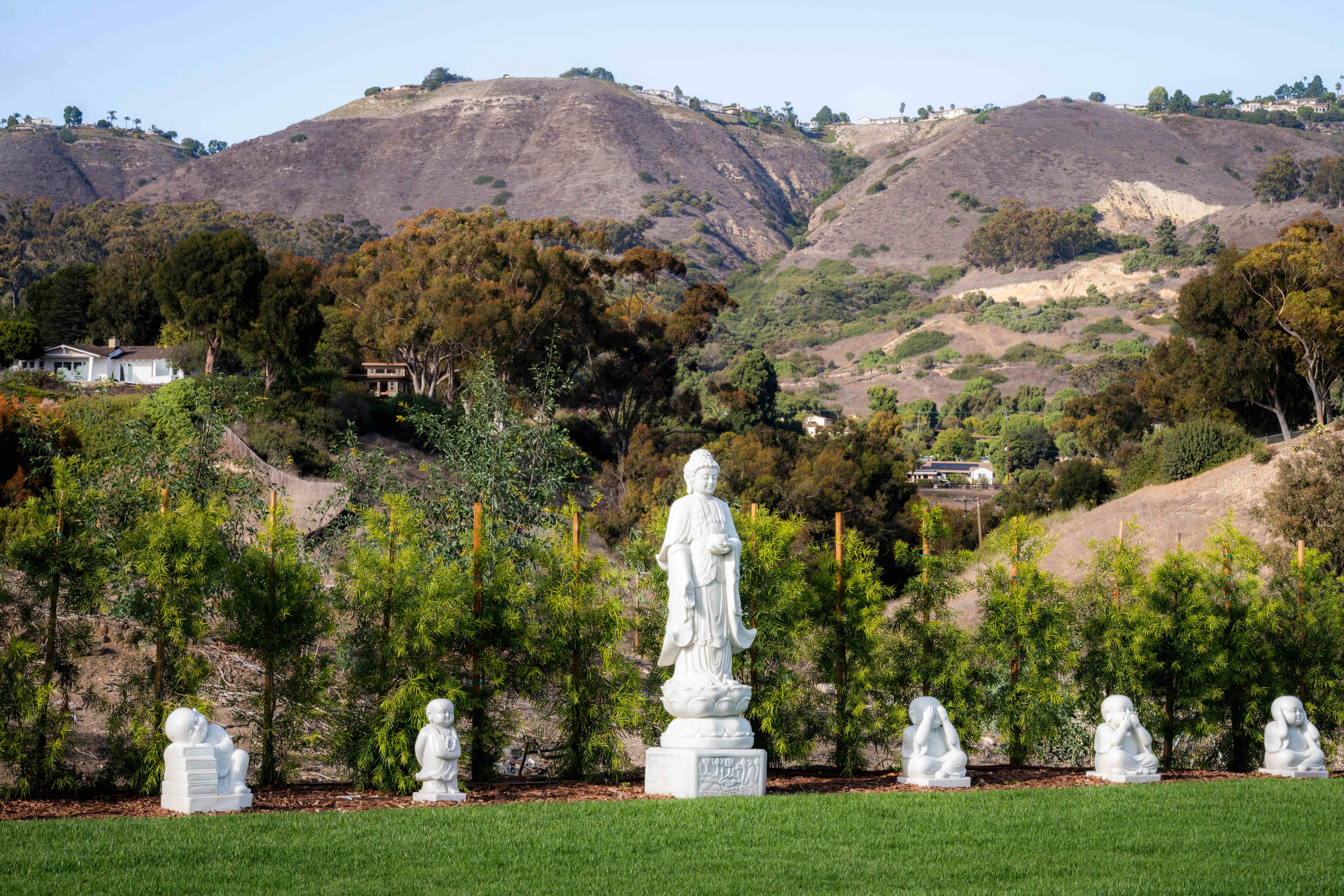 Standing Buddha statue with seated figures on lawn against rolling hills backdrop